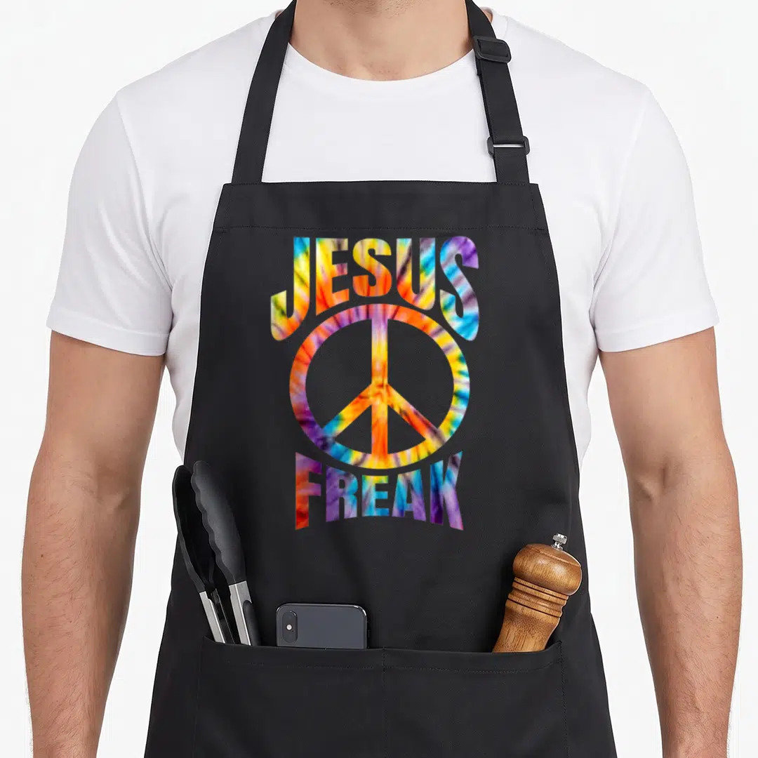 Jesus tie-dye apron - Arched JESUS FREAK text framing a peace symbol in red yellow blue purple tie-dye pattern — shown on a black cotton canvas apron against a white background