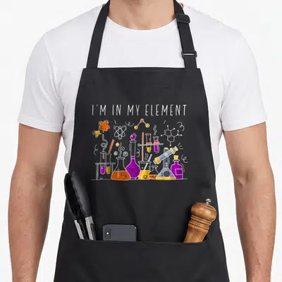 Chemist element chemistry apron - I'M IN MY ELEMENT text above cartoon flasks and molecules in purple orange yellow blue — shown on a black cotton canvas apron against a white background