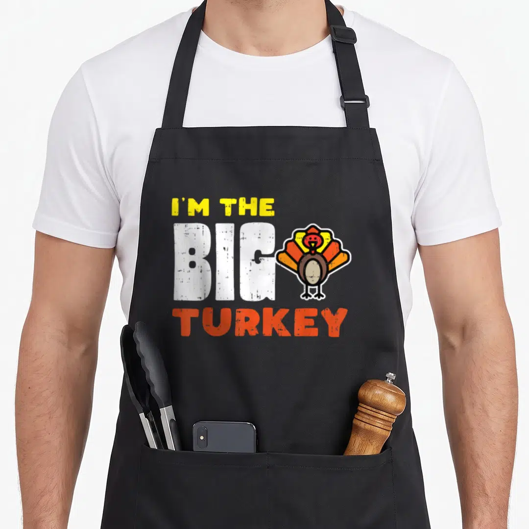 Thanksgiving family funny turkey apron - Stacked text reading I'M THE BIG TURKEY next to a cartoon turkey in yellow — shown on a black cotton canvas apron against a white background