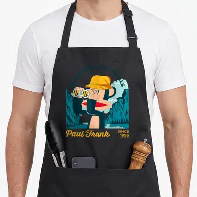 Paul black cat apron - Cartoon monkey in yellow hat with binoculars and SEARCHING FOR THRILLS text in blue and tan — shown on a black cotton canvas apron against a white background
