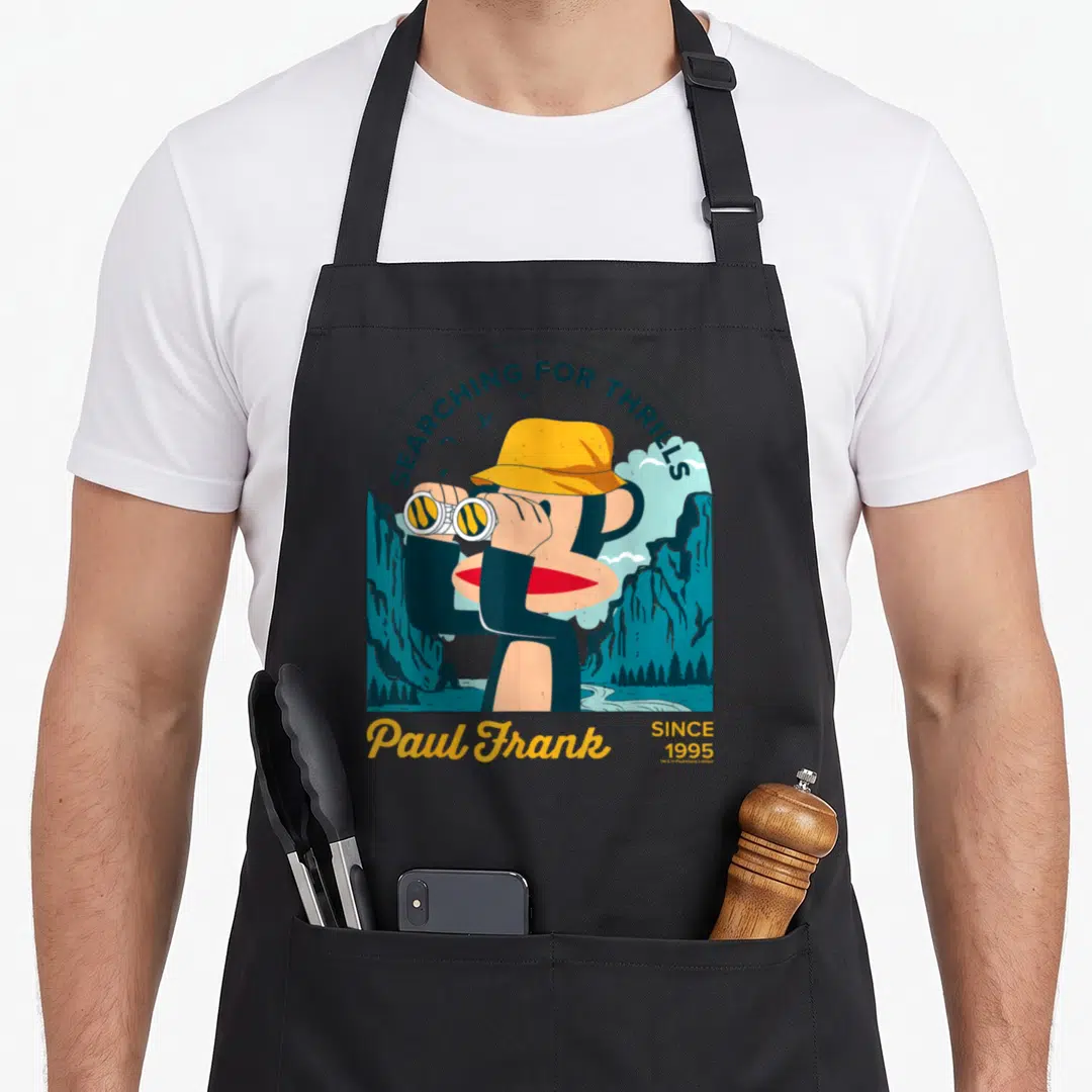Paul black cat apron - Cartoon monkey in yellow hat with binoculars and SEARCHING FOR THRILLS text in blue and tan — shown on a black cotton canvas apron against a white background
