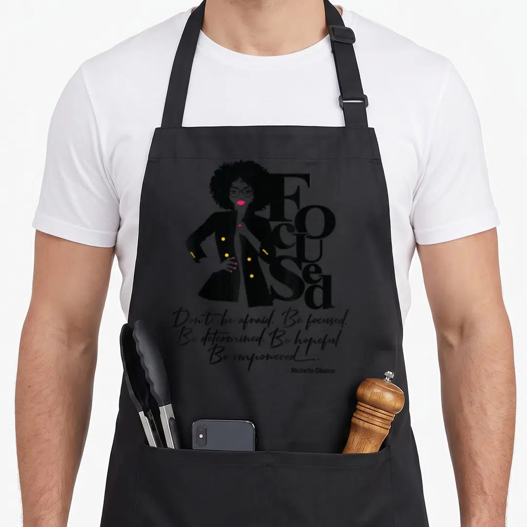 Diva magic apron - Stylized woman with afro and glasses overlapping Focused text and Michelle Obama quote — shown on a black cotton canvas apron against a white background
