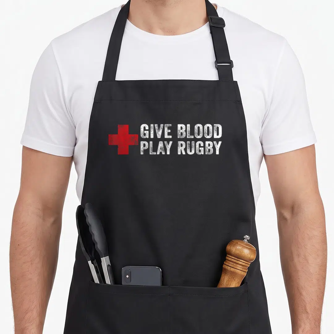 Give blood apron - Red cross symbol next to distressed white text reading GIVE BLOOD PLAY RUGBY — shown on a black cotton canvas apron against a white background