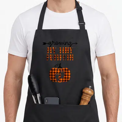 Tiny thanksgiving apron - Growing my tiny pumpkin text with orange plaid pumpkin containing white baby footprints — shown on a black cotton canvas apron against a white background