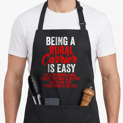 Rural letter carrier apron - White and red stacked text reading BEING A RURAL Carrier IS EASY except the bike is on fire — shown on a black cotton canvas apron against a white background