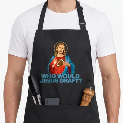 Would football apron - Cartoon Jesus holding brown football with blue text WHO WOULD JESUS DRAFT below — shown on a black cotton canvas apron against a white background