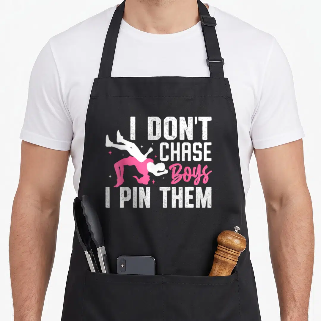 Wrestler wrestle wrestling apron - I Don't Chase Boys I Pin Them text with pink wrestler silhouette and stars — shown on a black cotton canvas apron against a white background