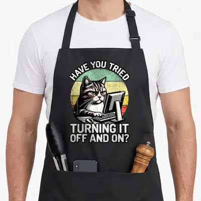 Tech cat apron - Grumpy cat at computer with retro sun and sarcastic tech support text in white black yellow green red — shown on a black cotton canvas apron against a white background
