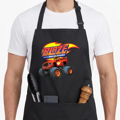 Monster machine apron - Red monster truck with blue eyes under Blaze logo and yellow flame graphic — shown on a black cotton canvas apron against a white background