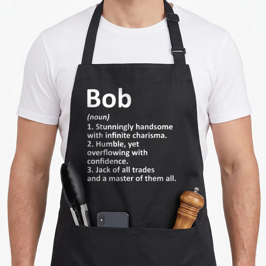 Definition birthday apron - White stacked text defining Bob as handsome, humble, and a master of trades — shown on a black cotton canvas apron against a white background