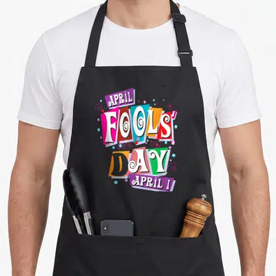 Fool prank apron - April Fools Day text on tilted colorful rectangles with spirals — shown on a black cotton canvas apron against a white background