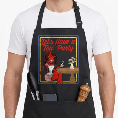 Horror tea party aprons - Red devil woman seated at table with teacup and flowers under Let's Have a Tea Party text — shown on a black cotton canvas apron against a white background