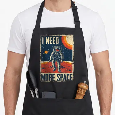 Cute astronaut space apron - Distressed astronaut on red planet with I Need More Space text in white orange red and blue — shown on a black cotton canvas apron against a white background