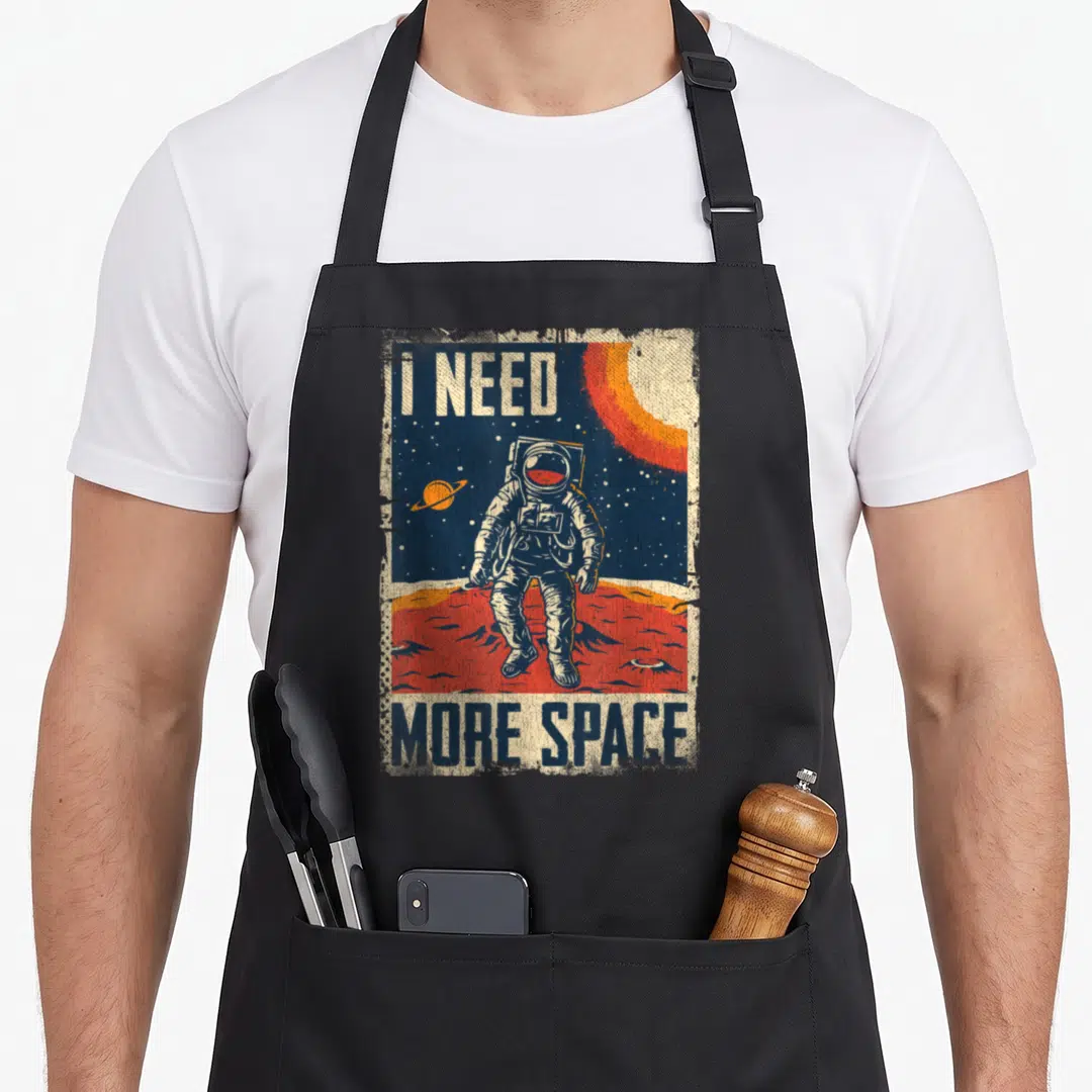 Cute astronaut space apron - Distressed astronaut on red planet with I Need More Space text in white orange red and blue — shown on a black cotton canvas apron against a white background