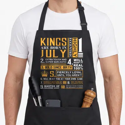 July king apron - Orange and white text listing Kings are born in July personality traits with crown icon — shown on a black cotton canvas apron against a white background