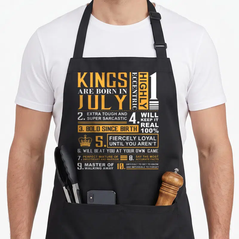 July king apron - Orange and white text listing Kings are born in July personality traits with crown icon — shown on a black cotton canvas apron against a white background