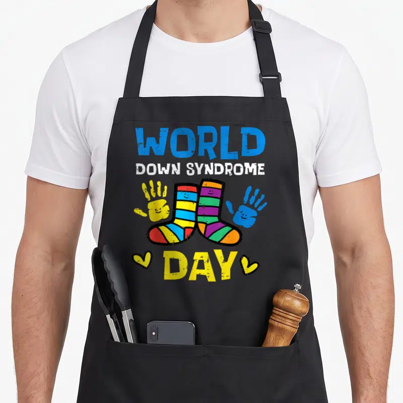 Syndrome teacher apron - Two striped socks with smiley faces flanked by yellow and blue handprints under text — shown on a black cotton canvas apron against a white background