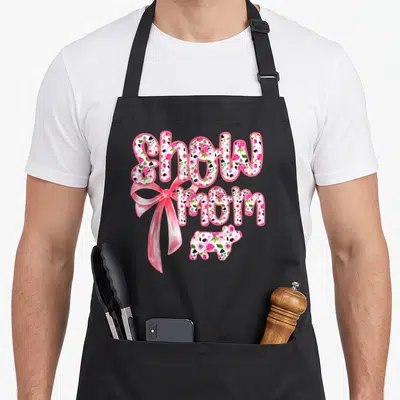 Coquette cooking mama apron - Show mom text with floral fill, pink ribbon, and small pig silhouette — shown on a black cotton canvas apron against a white background
