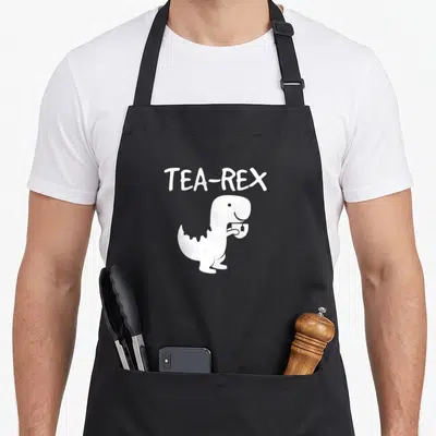 Tyrannosaurus tea party aprons - White TEA-REX text above cartoon dinosaur holding teacup — shown on a black cotton canvas apron against a white background