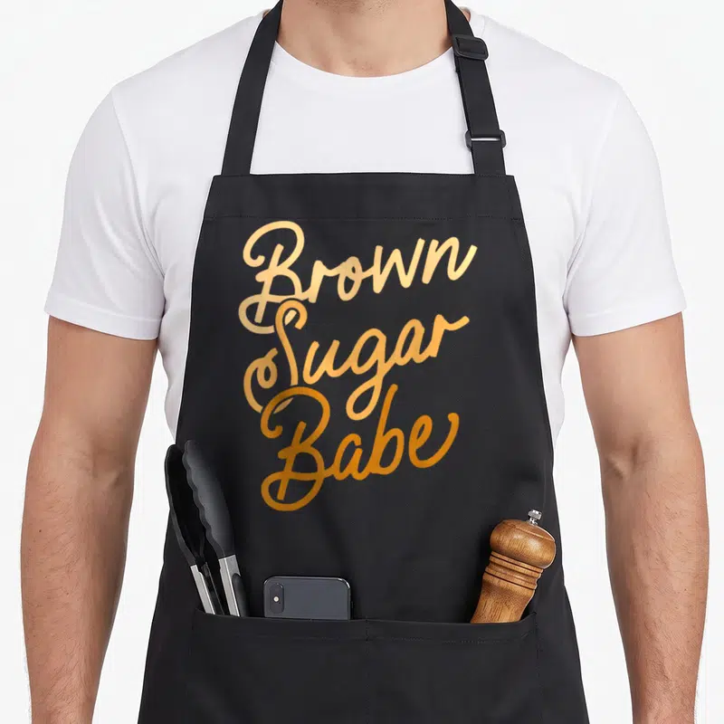Proud sugar apron - Orange script text reading Brown Sugar Babe stacked vertically on a dark background — shown on a black cotton canvas apron against a white background