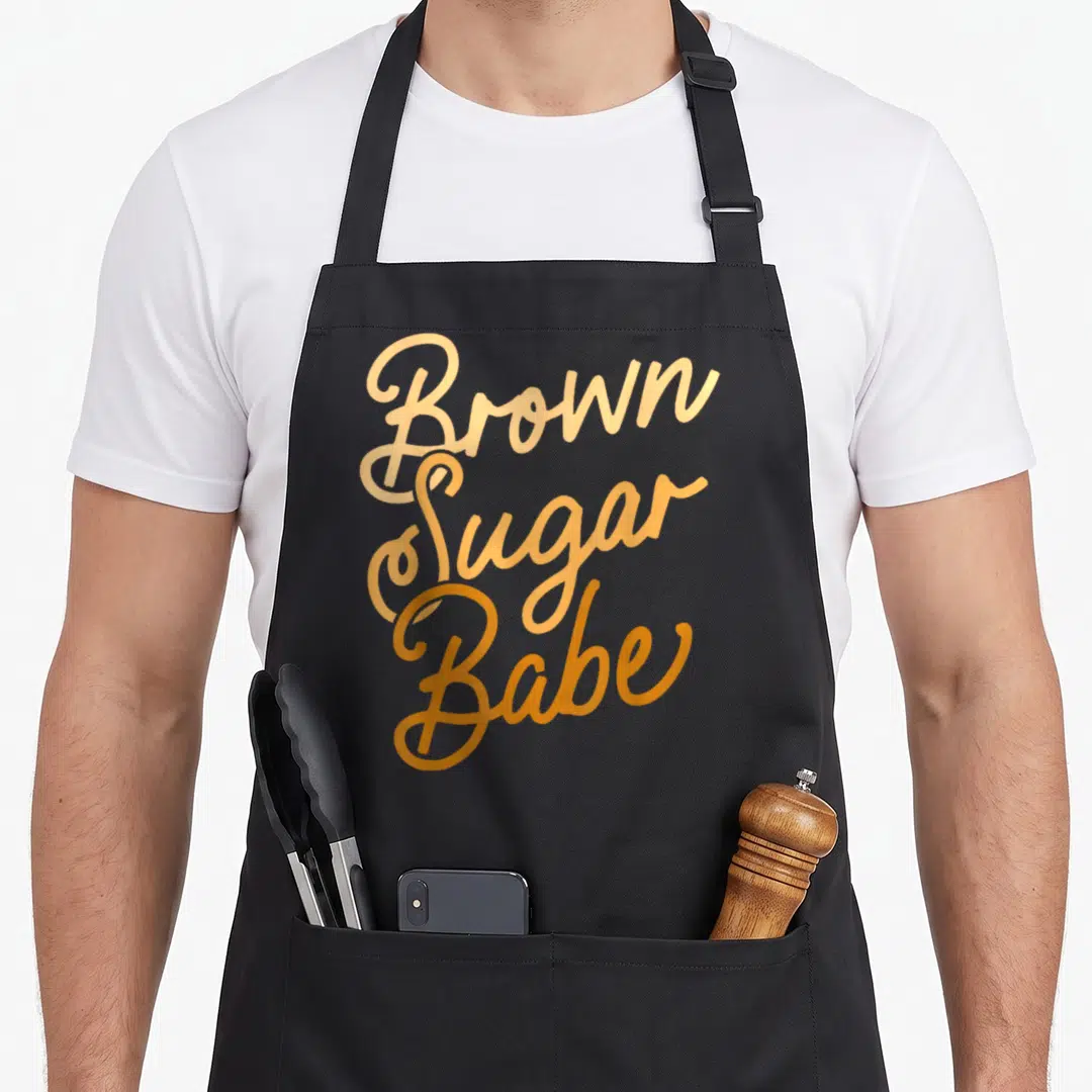 Proud sugar apron - Orange script text reading Brown Sugar Babe stacked vertically on a dark background — shown on a black cotton canvas apron against a white background