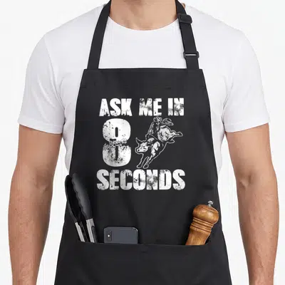 Ask cowboy apron - Distressed white ASK ME IN 8 SECONDS text with overlapping bull rider graphic — shown on a black cotton canvas apron against a white background