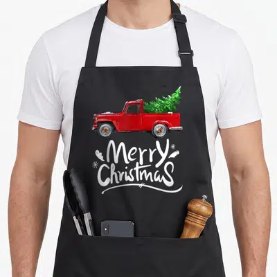 Wagon christmas tree apron - Red truck carrying green tree with white Merry Christmas text and snowflakes in distressed — shown on a black cotton canvas apron against a white background