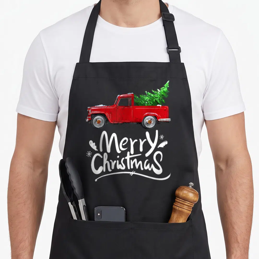 Wagon christmas tree apron - Red truck carrying green tree with white Merry Christmas text and snowflakes in distressed — shown on a black cotton canvas apron against a white background