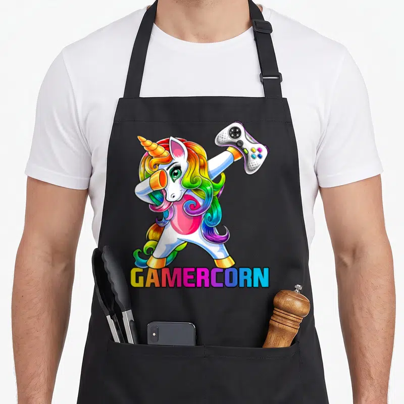 Gamercorn video game apron - Cartoon unicorn dabbing holding game controller with rainbow GAMERCORN text — shown on a black cotton canvas apron against a white background