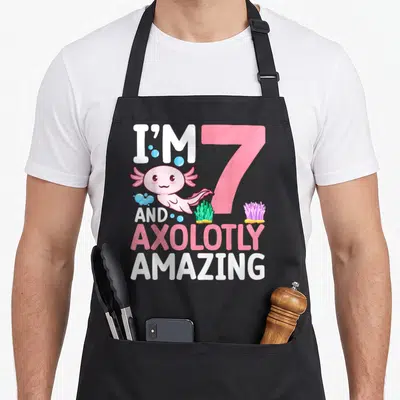 Axolotly birthday apron - Pink axolotl next to large 7 with bubbles, green seaweed, purple coral, and stacked text — shown on a black cotton canvas apron against a white background