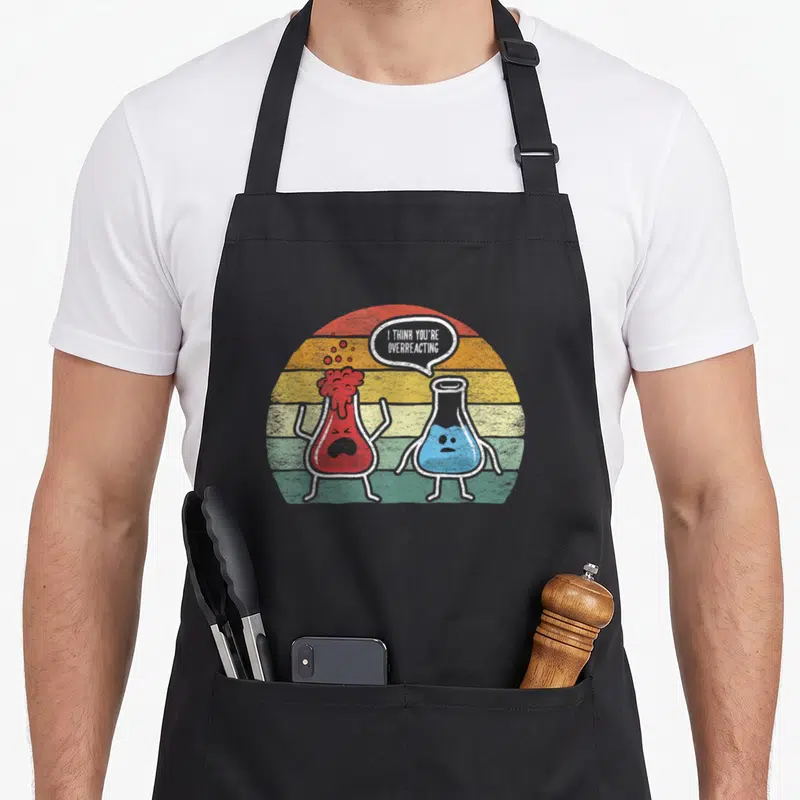 Overreacting nerd chemistry apron - Two cartoon beakers, red and blue, with speech bubble text I THINK YOU'RE — shown on a black cotton canvas apron against a white background