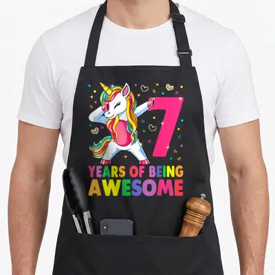 Unicorn dabbing birthday apron - Cartoon unicorn dabbing next to pink number 7 with rainbow mane and colorful text — shown on a black cotton canvas apron against a white background