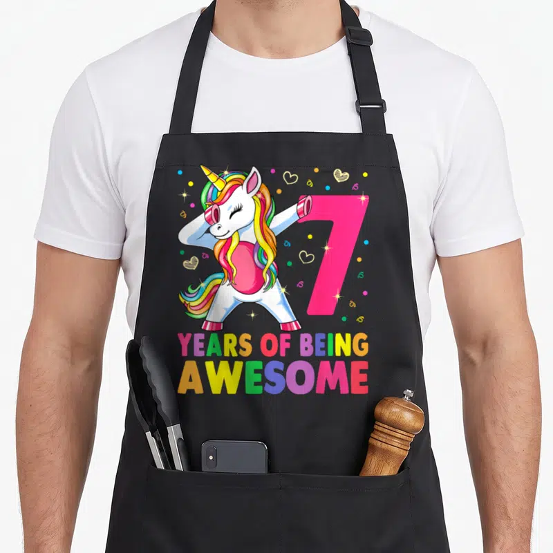 Unicorn dabbing birthday apron - Cartoon unicorn dabbing next to pink number 7 with rainbow mane and colorful text — shown on a black cotton canvas apron against a white background