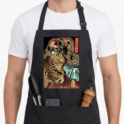 Fighting dragon apron - Japanese woodblock print of a tiger fighting a dragon with kanji text and blue water — shown on a black cotton canvas apron against a white background