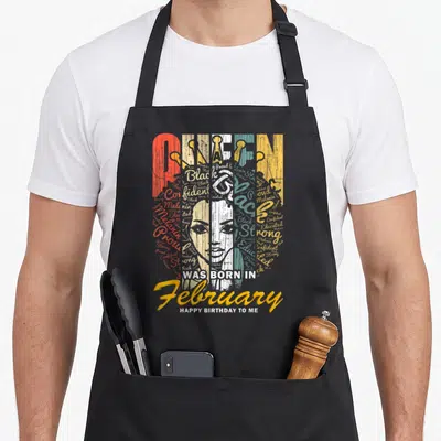 February queen apron - Typographic woman face made of words Confident Proud Strong Black Educated Melanin WAS BORN — shown on a black cotton canvas apron against a white background