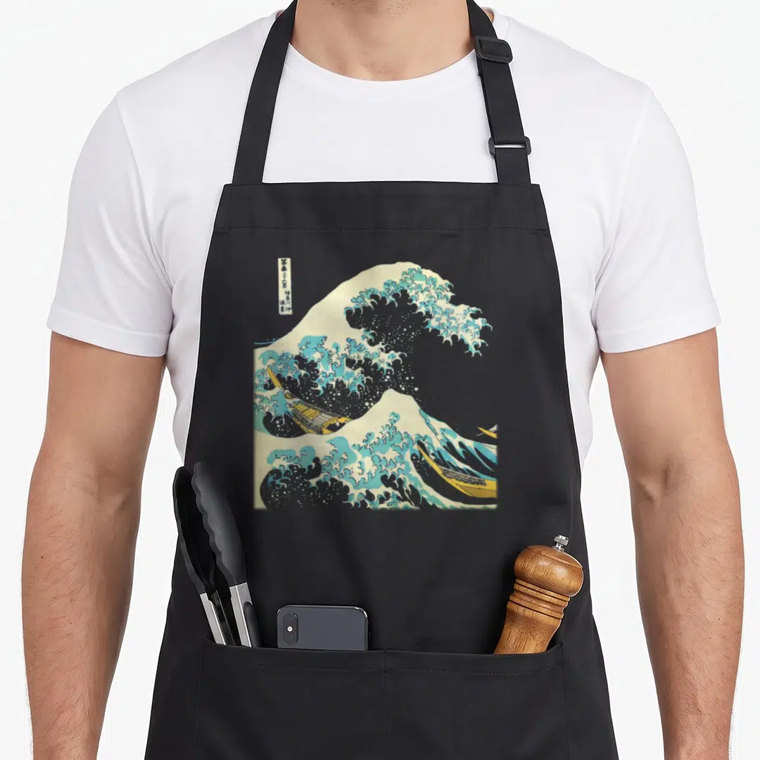 Hokusai wave apron - Blue and white wave with yellow boats and vertical Japanese text print — shown on a black cotton canvas apron against a white background