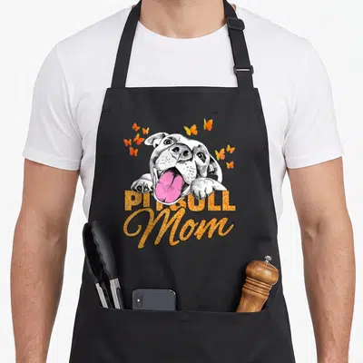 Butterfly dog mom apron - Cartoon pitbull head with pink tongue and orange butterflies surrounding PITBULL Mom text — shown on a black cotton canvas apron against a white background