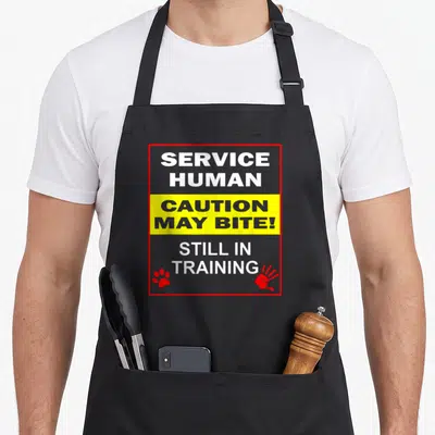 Service dog training apron - Warning sign graphic reading Service Human Caution May Bite Still In Training with paw — shown on a black cotton canvas apron against a white background
