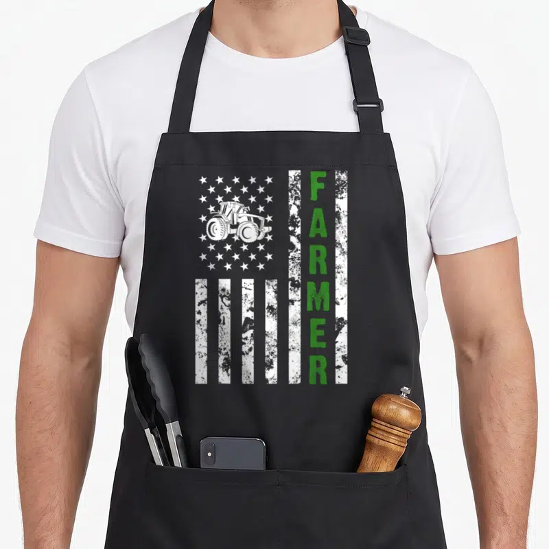 Farming farmer apron - Distressed flag design with white tractor in star field and vertical green FARMER text — shown on a black cotton canvas apron against a white background