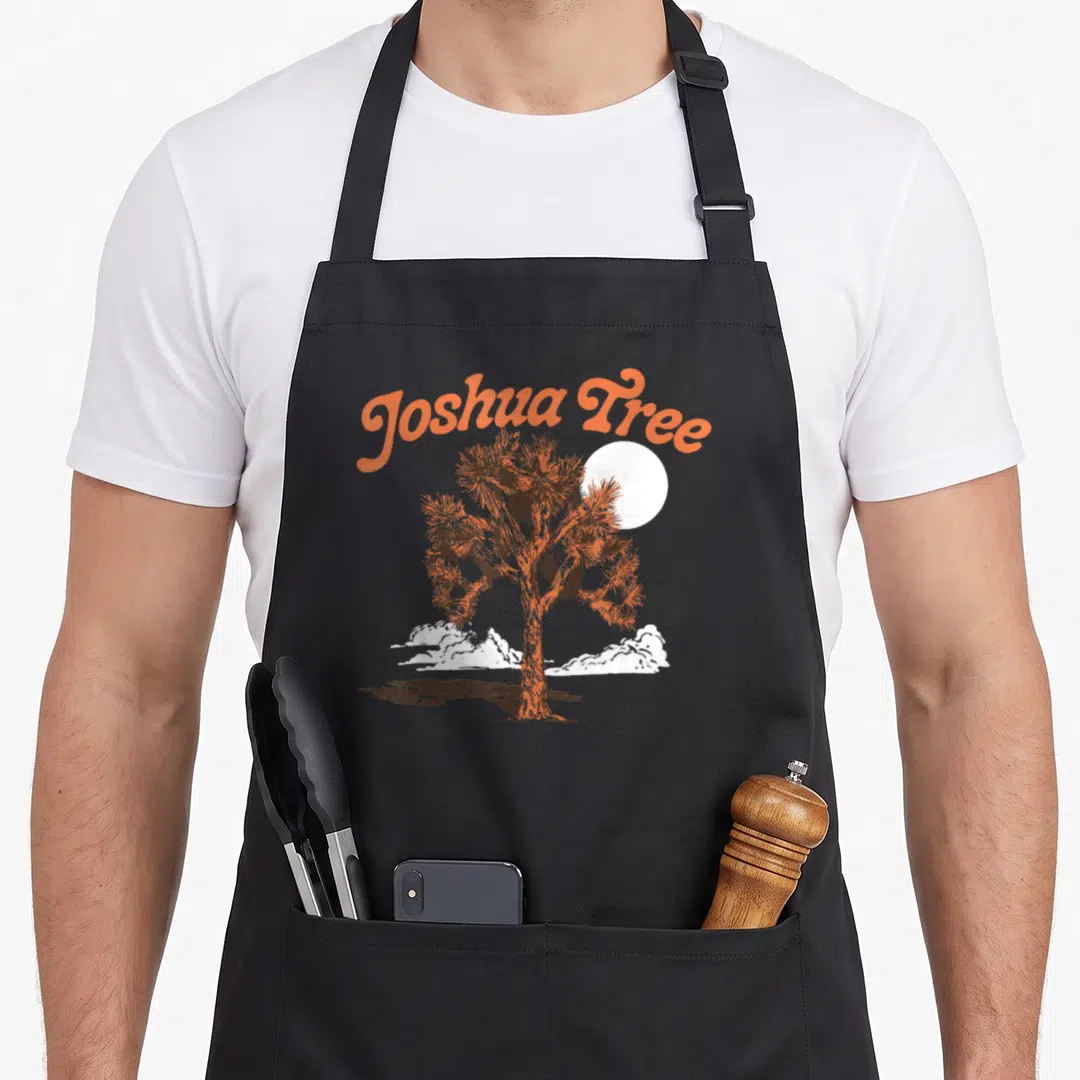 Eighty tree apron - Arched orange Joshua Tree text above brown tree, white sun and clouds — shown on a black cotton canvas apron against a white background