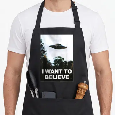 Alien hunter apron - UFO hovering over trees with I WANT TO BELIEVE text in white — shown on a black cotton canvas apron against a white background