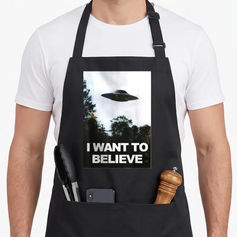 Alien hunter apron - UFO hovering over trees with I WANT TO BELIEVE text in white — shown on a black cotton canvas apron against a white background