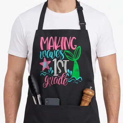 Mermaid making an apron - Pink and blue text reading MAKING waves IN 1ST grade next to a green mermaid tail — shown on a black cotton canvas apron against a white background