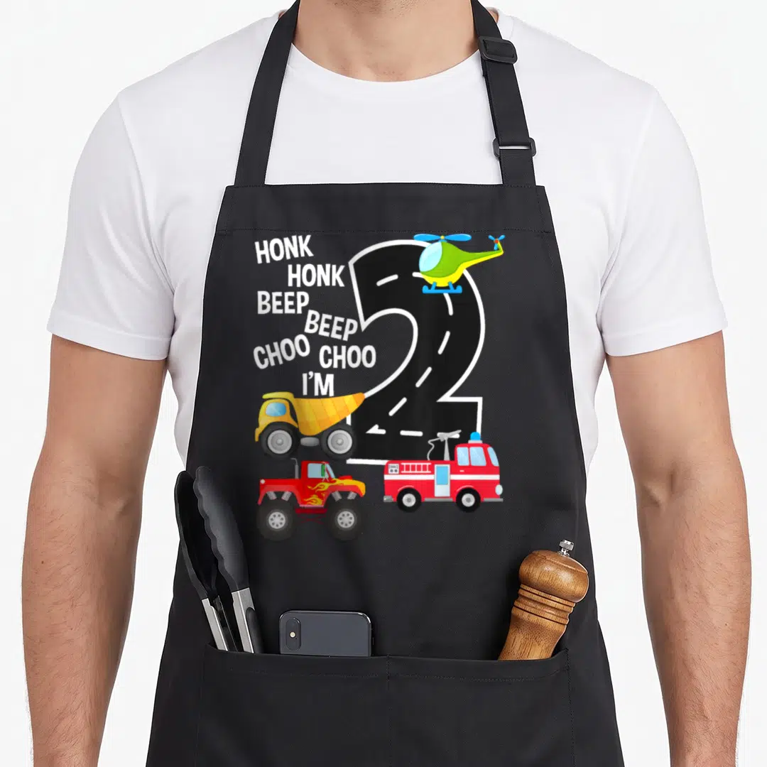 2nd truck apron - Road shaped number 2 with helicopter dump truck fire truck monster truck and honk beep choo text — shown on a black cotton canvas apron against a white background
