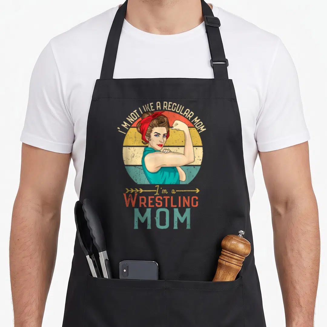 Gnd family aprons - Retro woman flexing arm in front of striped sun with Wrestling Mom text — shown on a black cotton canvas apron against a white background