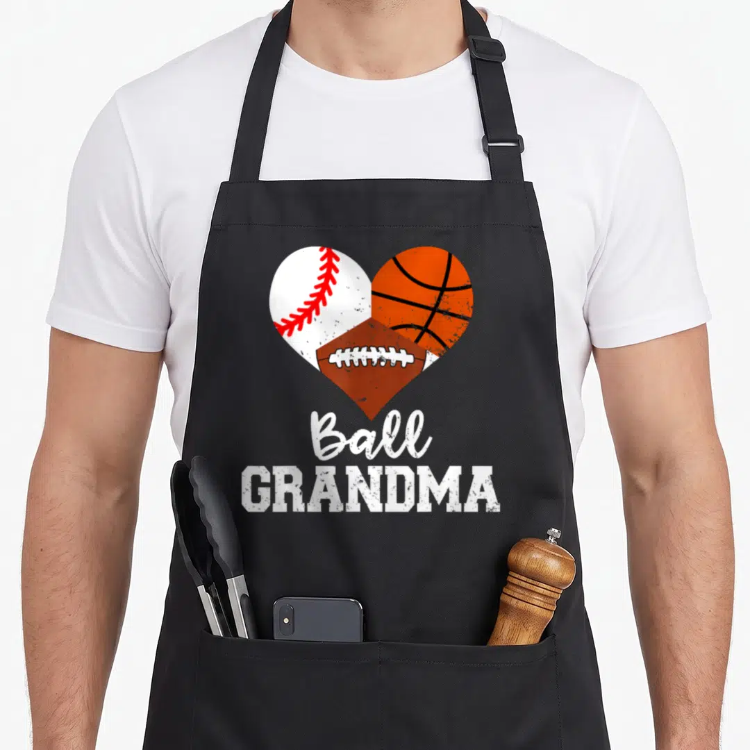 Ball grandma aprons - Heart shape formed by baseball, basketball, and football with stacked white text below — shown on a black cotton canvas apron against a white background