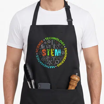 Stem student teacher apron - Circular STEM design with green yellow blue orange white science icons and text — shown on a black cotton canvas apron against a white background