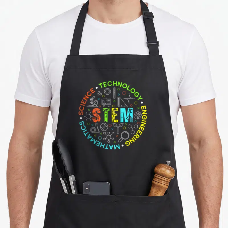 Stem student teacher apron - Circular STEM design with green yellow blue orange white science icons and text — shown on a black cotton canvas apron against a white background