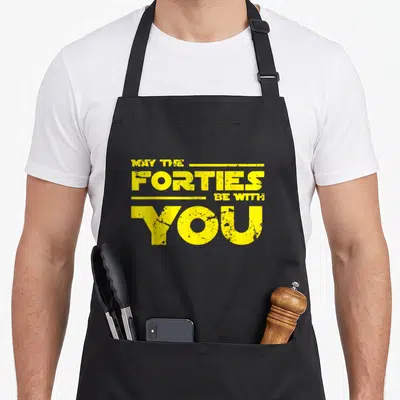 Forty 40th birthday apron - Yellow stacked text MAY THE FORTIES BE WITH YOU with horizontal lines — shown on a black cotton canvas apron against a white background