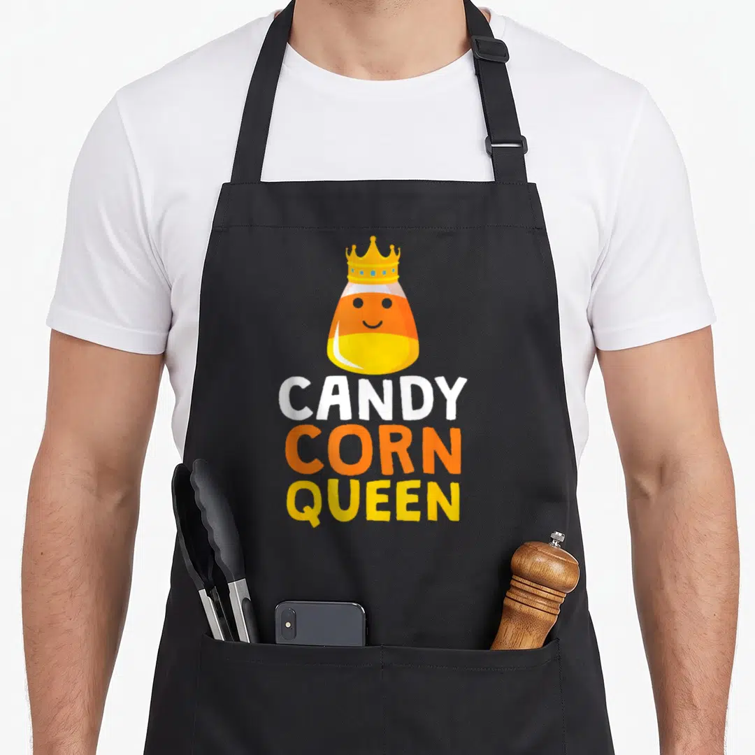 Queen halloween apron - Cartoon candy corn with crown and stacked CANDY CORN QUEEN text in yellow orange white — shown on a black cotton canvas apron against a white background